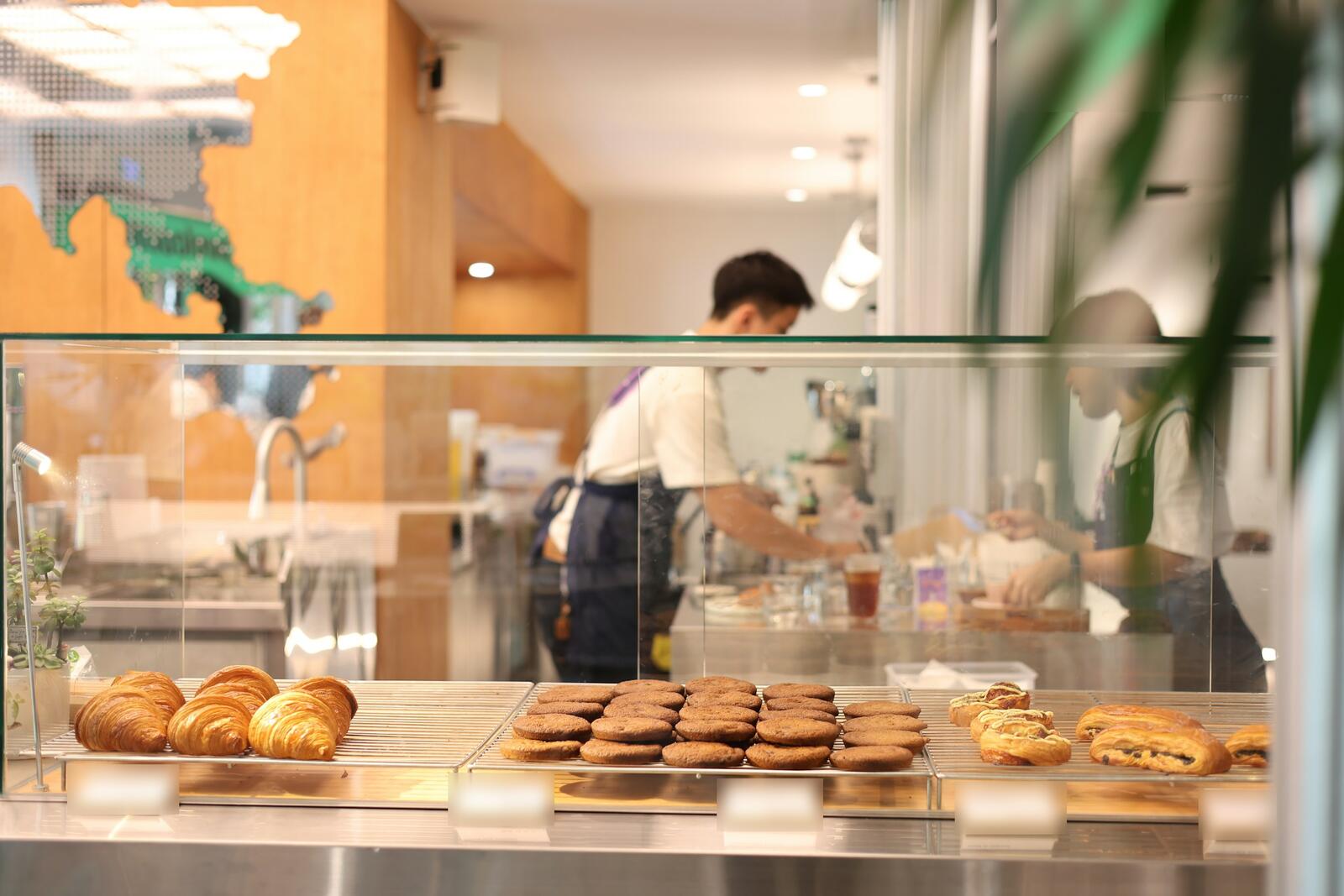 The bright bakery counter, with croissants and cookies under the glass and a baker at work behind it