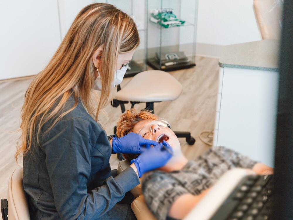 A hygienist in blue gloves cleaning a young patient's teeth in a bright, pale-wood room