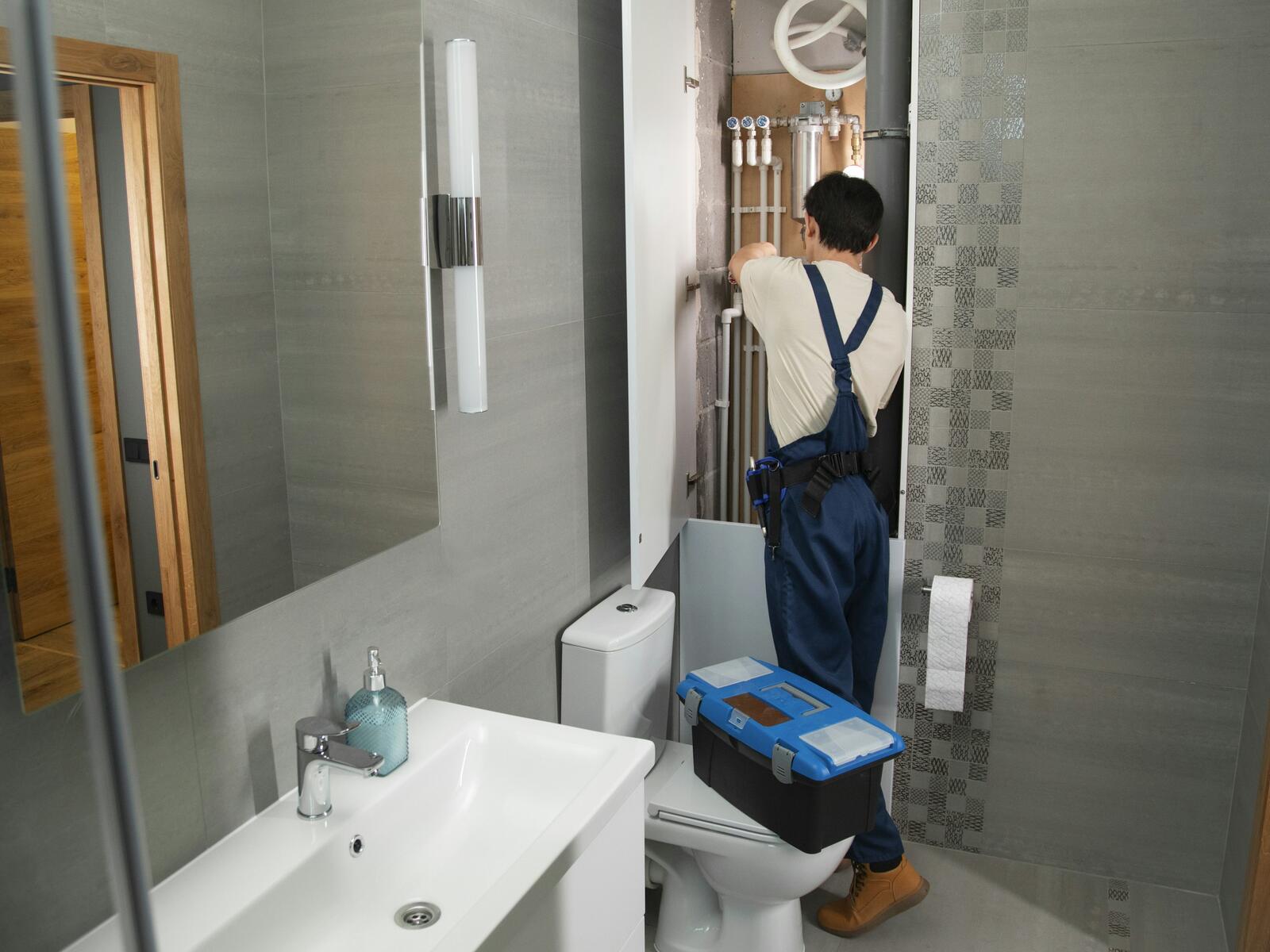 A plumber in blue overalls working on exposed wall pipes in a tiled bathroom