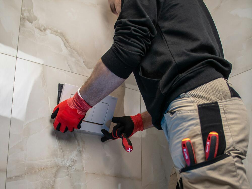 A plumber fitting a toilet flush plate on a marble-tiled bathroom wall