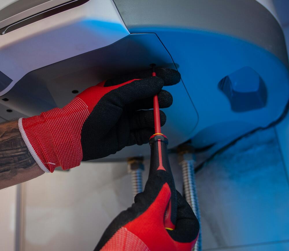 Gloved hands with a screwdriver working on the underside of a wall-mounted water heater